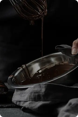 A hand holds a whisk dripping melted chocolate into a metal bowl covered with a gray cloth